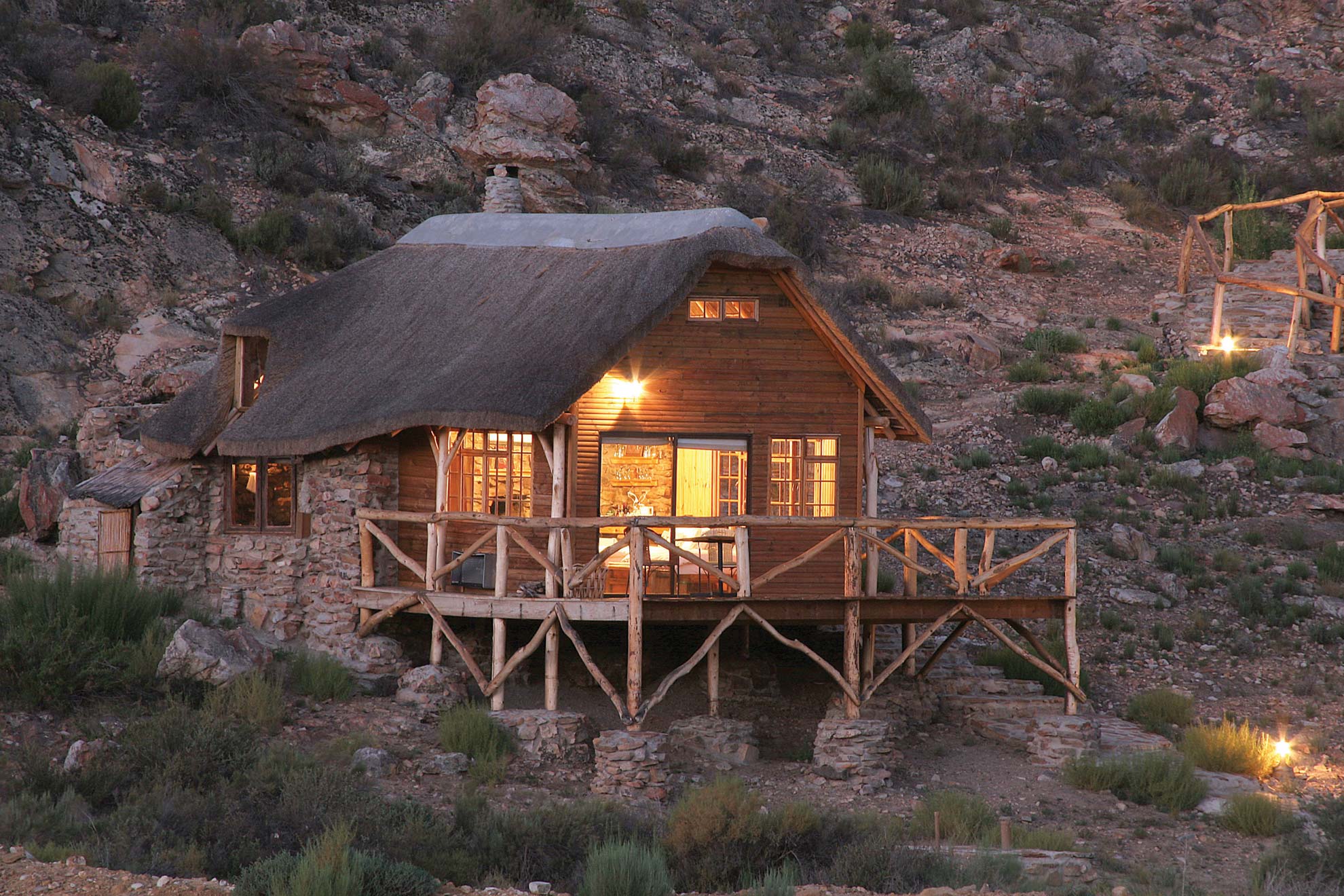 Premier Cottage, set against the rocky Karoo hillside, at night and with the lights on. Shown as part of Aquila's luxury overnight safari package.