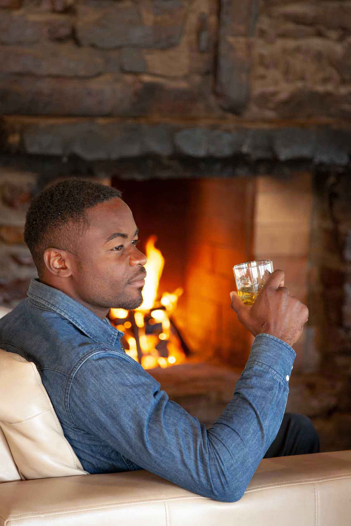 Man holding a tumblr glass of whiskey in front of a fireplace located by Aquila's indoor bar and overnight restaurant area.