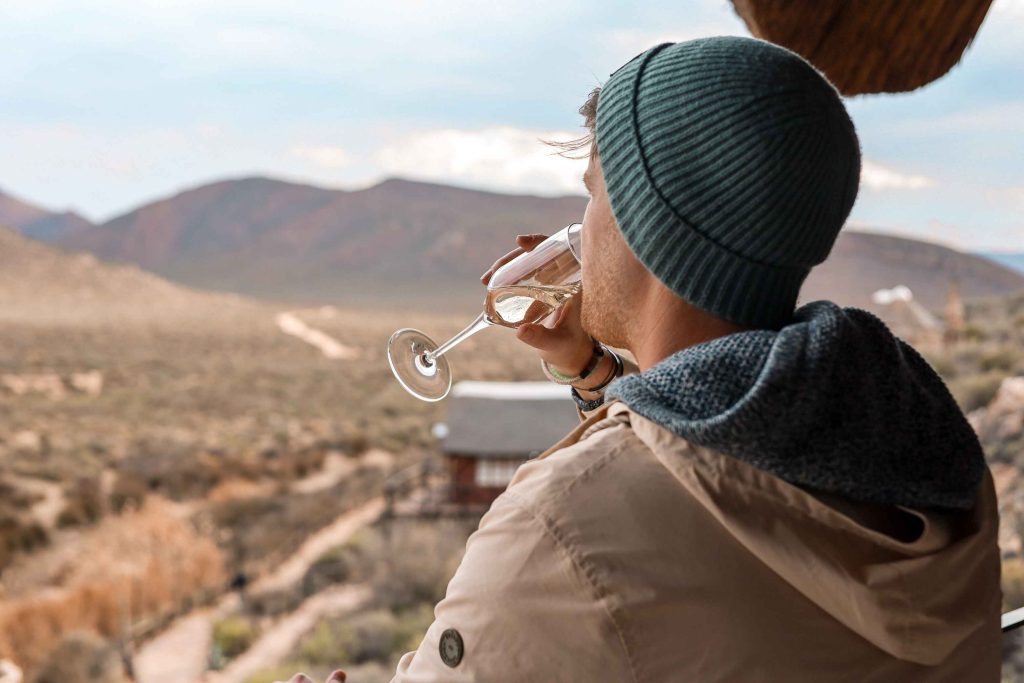 Man drinking champagne while overlooking the premier cottages at Aquila Private Game Reserve. Man is wearing a jacket and wool cap as it is a winter safari near Cape Town.